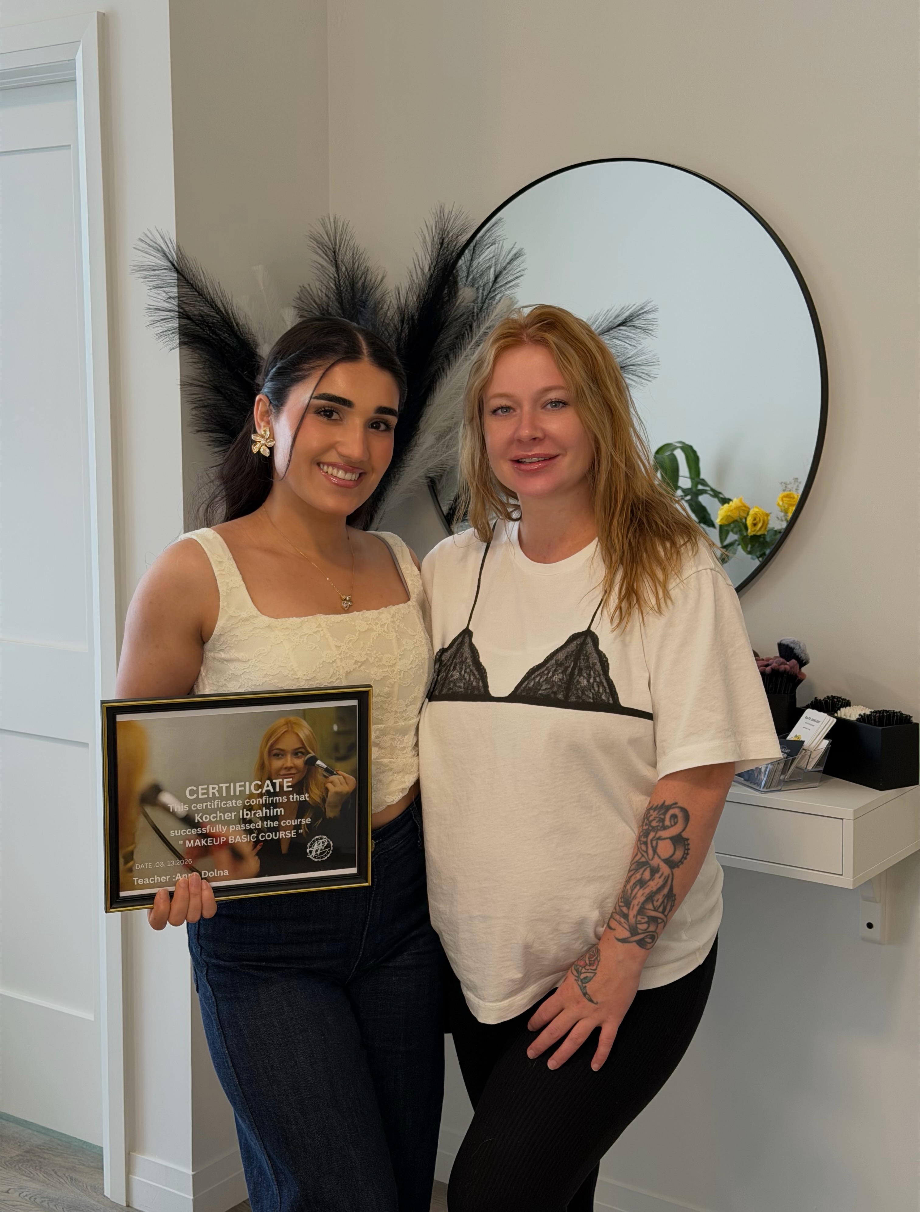 Kocer holding her makeup course certificate next to her teacher, the studio mirror behind them