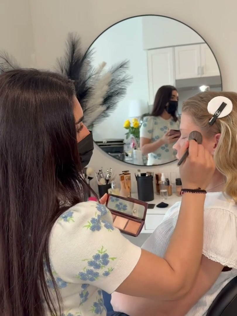 Kocer, masked, applying makeup at the round studio mirror, which reflects them both